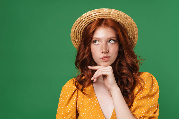 Image of ginger serious girl in straw hat thinking and looking aside isolated over green background - Foto, imagen