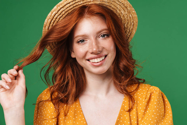 Obraz rudej radosnej dziewczyny w słomkowym kapeluszu uśmiechającej się i patrzącej na aparat odizolowany na zielonym tle - Zdjęcie, obraz