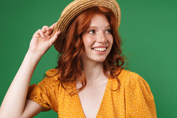 Image of ginger joyful girl in straw hat smiling and looking aside isolated over green background - Zdjęcie, obraz