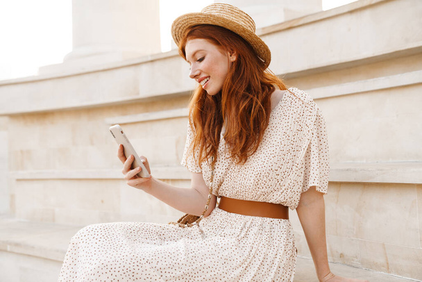Imagen de la joven pelirroja con sombrero de paja usando el teléfono móvil mientras está sentada en las escaleras en el día de verano - Foto, imagen