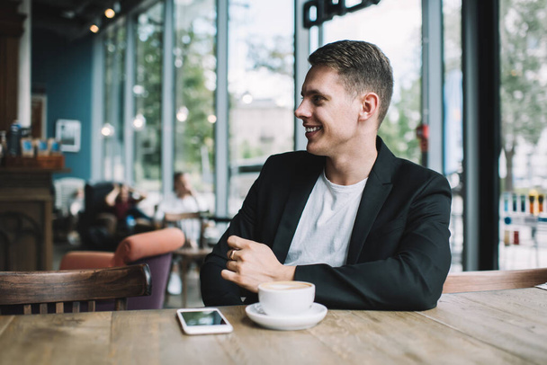 Positive young man in smart casual wear resting in cafe with full wall window smiling and looking away at daytime - Foto, imagen