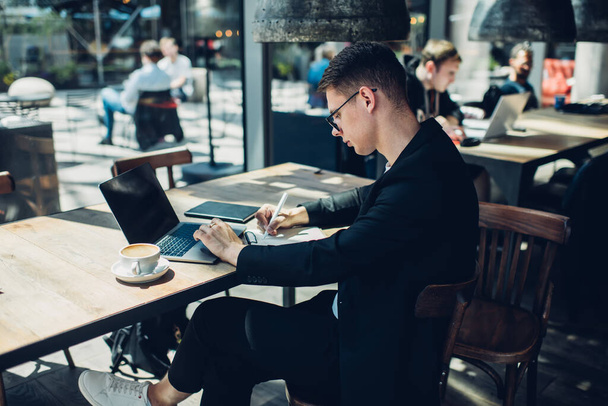Side view of young concentrated businessman writing notes and using laptop while working on project in modern cafe in daytime - Фото, изображение