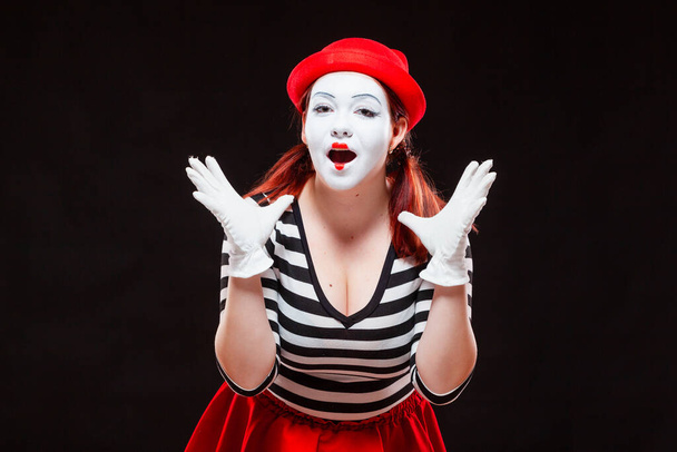 Portrait of female mime artist performing, isolated on black background. Woman in striped clothes and red skirt is standing with raised hands amazed. Symbol of wonder, admiration, worship - Foto, imagen