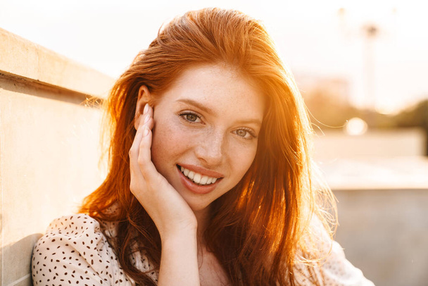 Image of cheerful ginger girl smiling and looking at camera at city street - Фото, изображение
