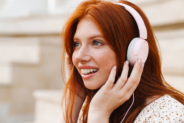 Image of cheerful ginger girl smiling and listening music with headphones at city street - Zdjęcie, obraz