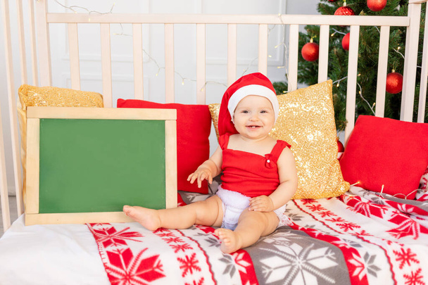 happy little baby six months in a Santa hat sitting in a colb at home by the Christmas tree with a text Board, New year and holiday concept - Fotografie, Obrázek