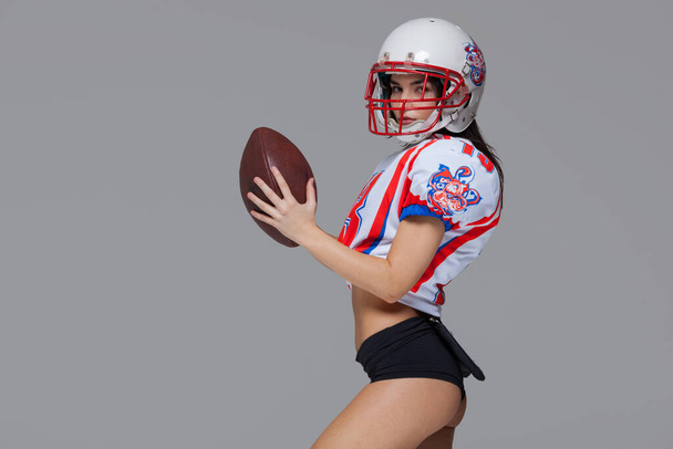 Sporty girl wearing sexy uniform of American football player and helmet posing with ball ready to throw isolated on grey background - Fotoğraf, Görsel