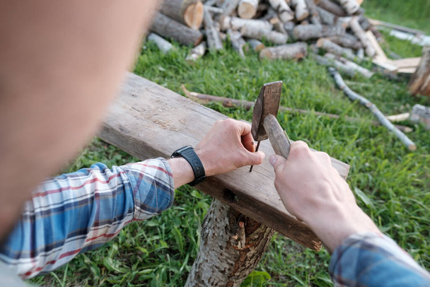 Τα αρσενικά χέρια ενός ξυλουργού φτιάχνουν ένα παγκάκι σφυρηλατώντας καρφιά σε μια σανίδα σε μια αυλή του χωριού. Άνω όψη. - Φωτογραφία, εικόνα