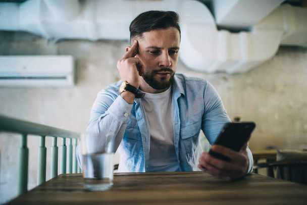 Serious bearded man in casual clothes sitting at wooden table with glass of water in cafe and concentrating while using smartphone - Photo, Image