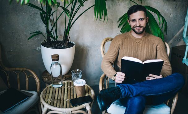 Bearded male sitting on armchair and reading interesting book in modern cafeteria wearing casual clothes smiling and looking at camera - Foto, immagini