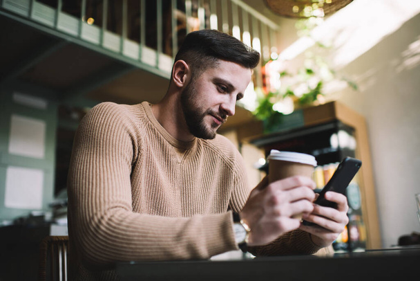 Smiling black haired bearded man in casual clothes holding cup of coffee and browsing smartphone while resting in cozy cafe - Photo, Image