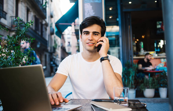 Young handsome man is casual clothes using laptop and talking on smartphone while sitting in cafe in Barcelona city and looking at camera - Foto, Imagem