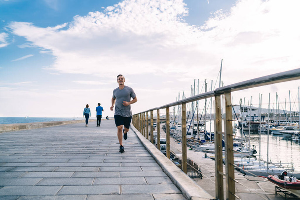 Smiling caucasian male jogger enjoying training on coastline feeling energy and vitality for keeping perfect body shape, 20s cheerful young man running on street on morning cardio workout - Foto, afbeelding