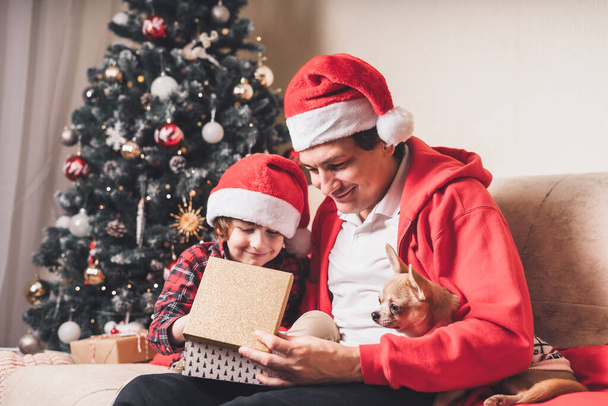 Happy family in Santa hat, father and child boy open Christmas gift at home. Sitting on a couch in the living room with puppy dog in sweater - Fotó, kép