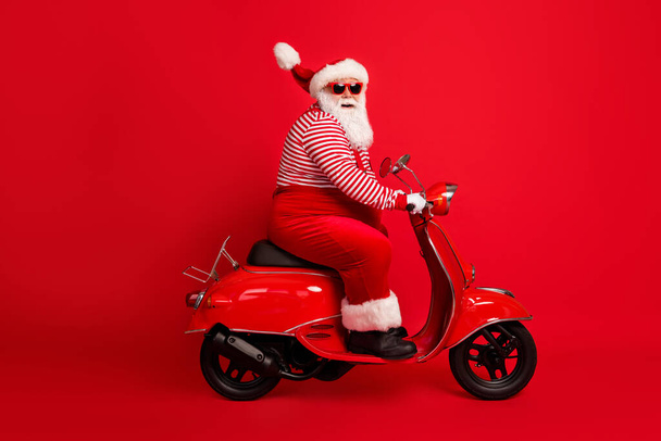Full length profile photo of grandfather grey beard drive retro scooter look camera wear santa claus x-mas costume suspenders sunglass striped shirt cap isolated red color background - Fotografie, Obrázek