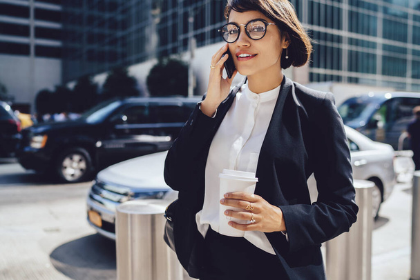 Confident young woman in classy clothes and glasses with cup of hot drink talking on cellphone while walking down busy street and looking at camera in New York City - Φωτογραφία, εικόνα