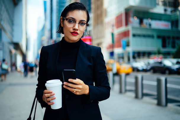 Elegant young woman in formal wear and eyeglasses holding cup of hot drink and looking at camera while standing in modern city centre - Foto, Imagen