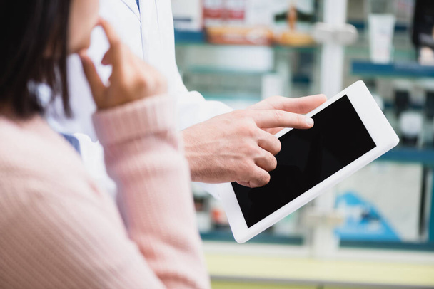 przycięty widok farmaceuty wskazujący palcem na cyfrową tabletkę z pustym ekranem w pobliżu klienta  - Zdjęcie, obraz