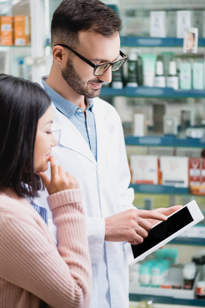 farmacista in occhiali puntando con il dito al tablet digitale con schermo bianco vicino cliente asiatico  - Foto, immagini