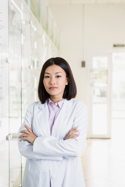 brunetka azjatycki farmaceuta w biały płaszcz stojący z krzyżowane ramiona w apteka - Zdjęcie, obraz