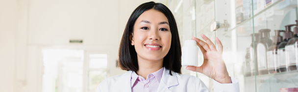 cheerful asian pharmacist in white coat holding bottle in drugstore, banner - Photo, Image