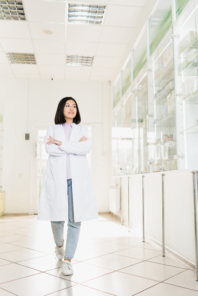 täyspitkä hymyilevä aasialainen farmaseutti valkoisessa takissa kävely ristissä kädet apteekissa - Valokuva, kuva