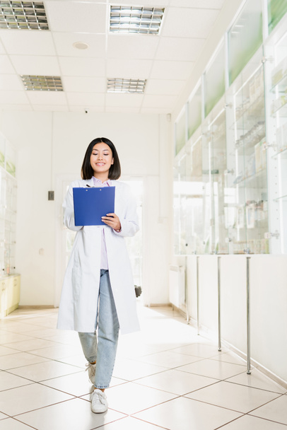 full length of cheerful asian pharmacist in white coat holding clipboard in drugstore  - Foto, afbeelding