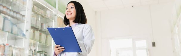cheerful asian pharmacist in white coat holding clipboard in drugstore, banner - Photo, image
