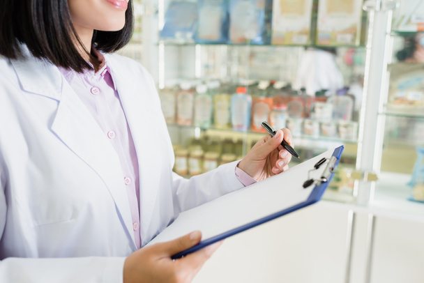 bijgesneden mening van apotheker in witte jas houden pen in de buurt van klembord in drogisterij  - Foto, afbeelding