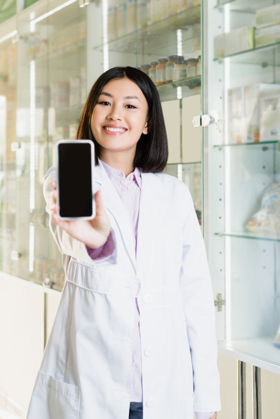 allegro farmacista asiatico in bianco appendiabiti smartphone con schermo bianco in primo piano sfocato  - Foto, immagini