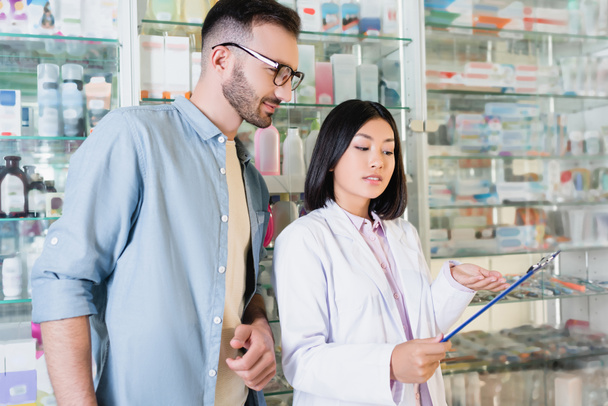 веселый азиатский фармацевт в белом халате, указывающий рукой на планшет возле человека в аптеке  - Фото, изображение