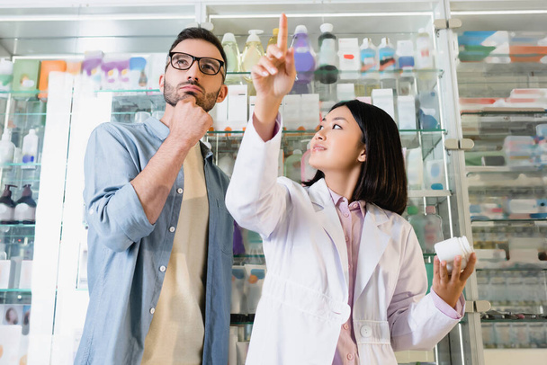 Низкий угол зрения азиатского фармацевта указывая пальцем и глядя на задумчивого клиента в очках в аптеке - Фото, изображение