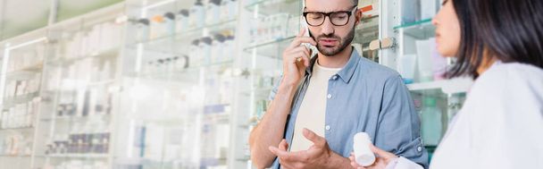 customer in eyeglasses talking on smartphone near asian pharmacist with bottle of medication on blurred foreground, banner - Fotó, kép