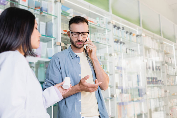 client en lunettes parlant sur smartphone près d'un pharmacien asiatique avec bouteille de médicament au premier plan flou - Photo, image