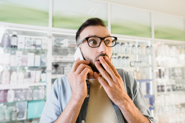 Geschockter Mann mit Brille spricht auf Smartphone und verdeckt Mund in Drogerie - Foto, Bild