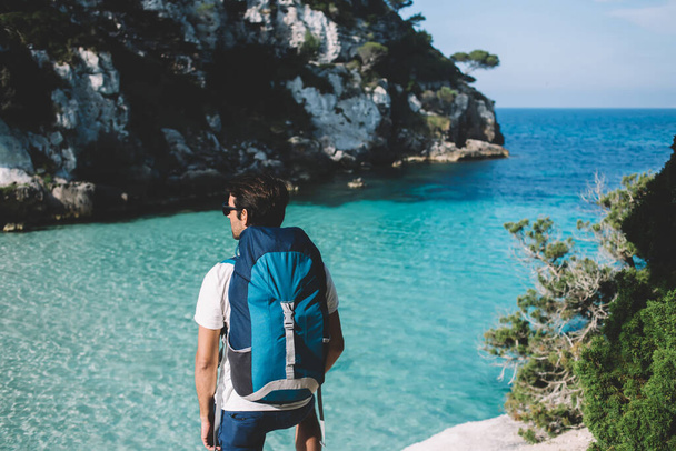 Back view of young man tourist with travel backpack enjoying beauty of seashore, experienced male hiker looking at natural scenic landscape while spending time for exploring Spanish Menorca island - Photo, Image