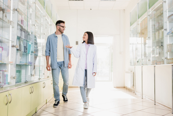 полная длина веселый азиатский фармацевт в белом халате ходьба с клиентом в аптеке - Фото, изображение
