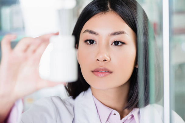 Азиатский фармацевт в белом халате смотрит на бутылку на размытом переднем плане - Фото, изображение