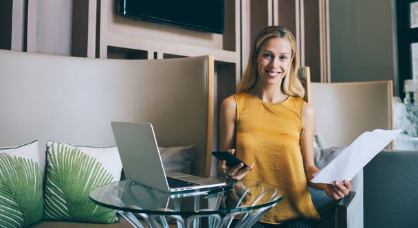 Delighted businesswoman in casual outfit sitting with documents and gadgets at table and  working remotely in cafe while smiling and looking at camera - 写真・画像