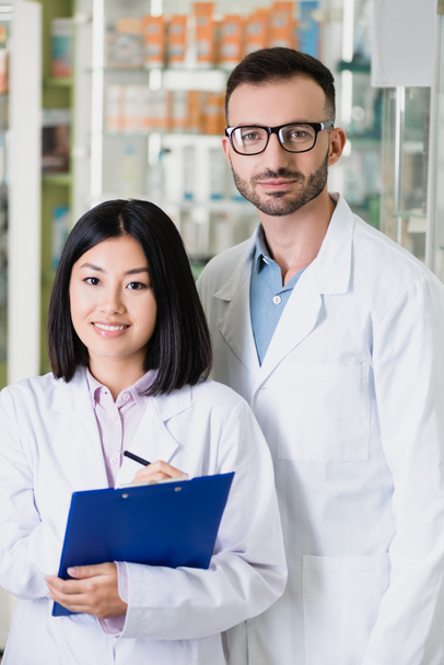 pharmaciens interracial souriants en manteaux blancs regardant la caméra en pharmacie - Photo, image