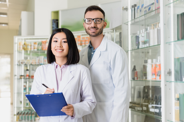 farmacéuticos interracial feliz en batas blancas mirando a la cámara en la farmacia - Foto, Imagen