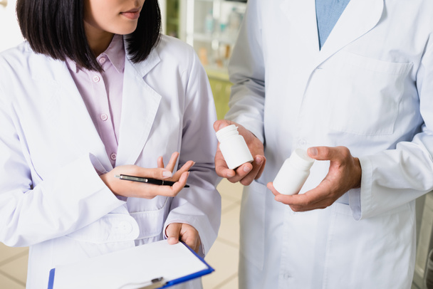 częściowy widok farmaceuty w białym fartuchu trzymającego butelki z lekami w pobliżu kolegi w aptece - Zdjęcie, obraz