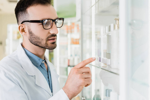bearded pharmacist in white coat and eyeglasses pointing with finger in drugstore  - Fotó, kép