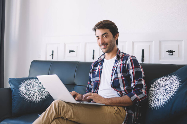 Confident ethnic male freelancer in stylish casual wear sitting on sofa with crossed legs smiling and looking at camera while working with laptop in Barcelona - Foto, Imagen