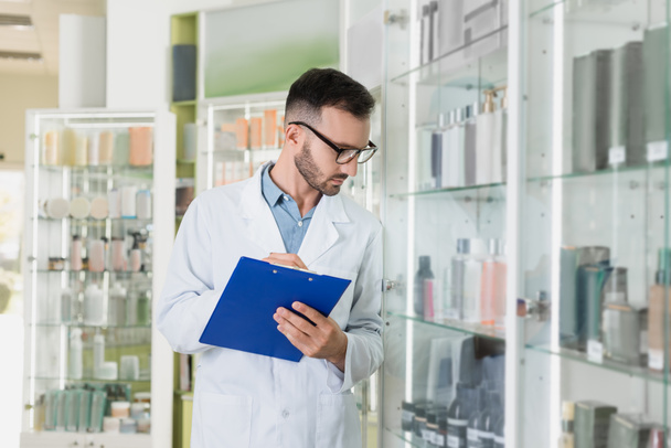 bearded pharmacist in white coat and eyeglasses holding clipboard while checking medication in drugstore  - Foto, Bild