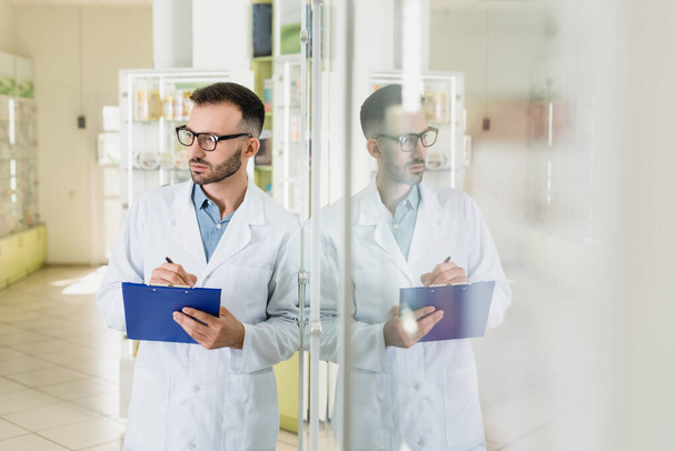szakállas gyógyszerész szemüveg gazdaság vágólapon, miközben ellenőrzi a gyógyszert a gyógyszertárban  - Fotó, kép