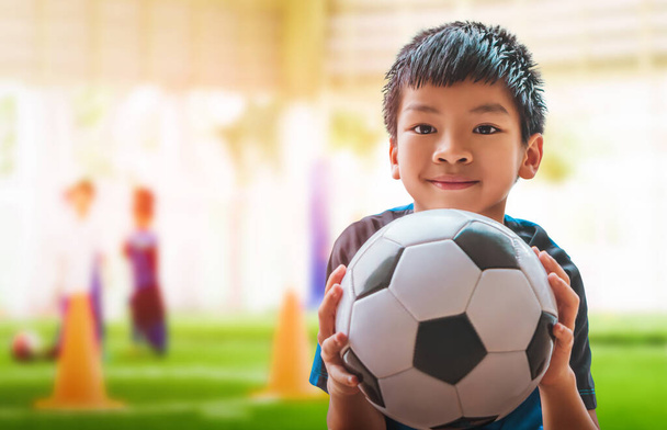 Asiatischer kleiner Fußballjunge mit einem Lächeln hält einen Fußballball mit Trainingsplatz im Hintergrund. - Foto, Bild