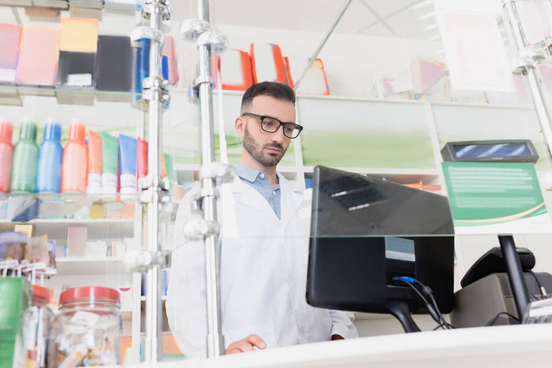 pharmacist in white coat and eyeglasses looking at computer monitor in drugstore - Fotó, kép