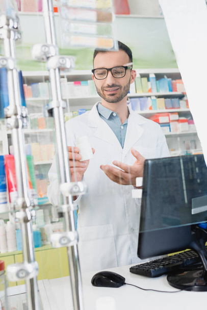 šťastný lékárník v bílém plášti a brýlích ukazuje rukou na láhev poblíž monitoru počítače v lékárně - Fotografie, Obrázek