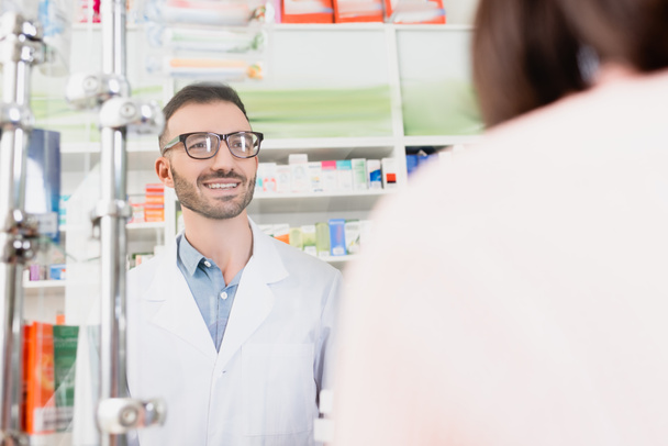 smiling pharmacist in white coat and eyeglasses looking at customer on blurred foreground - Foto, Imagen